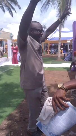 A man in a grey outfit and sandals is dancing at an outdoor event, likely a wedding, on a partially grassy and dirt surface. He is holding a blue object, possibly a brochure or program, in his hands. He has his arms raised and is swaying his body to the music. Other people are visible in the background, including a person holding a microphone and another man in a patterned shirt walking away. Tents, palm trees, and decorations suggest an organized celebration. The weather appears sunny.
