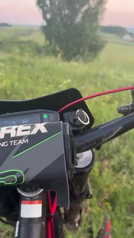 The video opens with a close-up of a dirt bike's handlebars. The digital display shows "N" for neutral, and a key is inserted into the ignition. A red wire is visible, running from the handlebars upwards. The background is a grassy field with scattered wildflowers. As the video progresses, the camera pans to the right, revealing more of the handlebars, including a mirror reflecting the environment and a "ProTaper" grip. A hand wearing a black and white motocross glove is then shown gripping the handlebar. The backdrop remains the same grassy field, with a gentle slope leading down to a distant village under a pastel-colored sky. The sun appears to be setting, casting a soft light over the landscape. The video ends with the camera focused on the gloved hand on the handlebar.