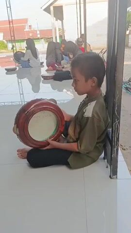 A boy sits on the ground, holding a round drum with his left hand and resting his right elbow on it. He is wearing a green and white shirt and black pants. The drum has a white surface with some writing on it. In the background, other people, including young women and children, are seated on the floor, some of them also holding drums. The scene appears to be outdoors, with a building and trees visible in the distance. The lighting suggests it is daytime.