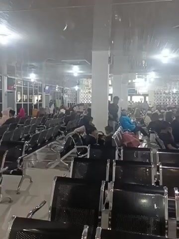 A wide shot shows a crowded waiting area with rows of empty black chairs in the foreground and people seated in chairs further back. The room is well-lit by fluorescent lights and appears to be a public space like a waiting room or terminal. The people are diverse in age and gender, many wearing masks. Some are looking at their phones, others are talking, and some appear to be sleeping. The atmosphere seems calm, despite the crowd.