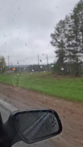 From the perspective of inside a car, the video shows a countryside scene on a rainy day. The car is moving on a wet dirt road.  Outside the car window, there is a green field, a line of wooden power poles, and trees lining the edge of the field and extending into the distance. Raindrops are visible on the car window and side mirror. The sky is overcast.