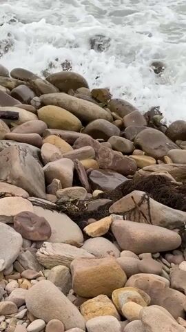 The video shows a rocky shoreline with waves crashing onto the stones. The rocks are of various sizes and colors, ranging from light gray to dark brown. Some of the rocks are wet from the waves, and there is some seaweed visible amongst the stones. The water is choppy and white with foam. The camera is positioned low, close to the ground, looking up towards the sea. The lighting is overcast, suggesting a cloudy day.
