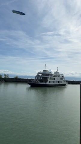 A ferry boat is seen sailing on the water. Above the ferry, a blimp is flying in the sky. The sky is blue with some clouds. The water is calm and appears to be a lake or a bay. There is a pier in the background with some trees on the land behind it. The blimp has writing and a logo on it.