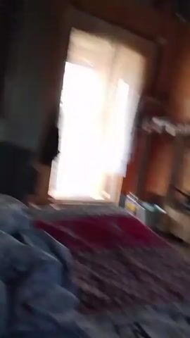 The video shows a messy room with a bed in the foreground covered with a patterned blanket. There is a window with curtains on the left, and the room has wooden walls. Various objects are scattered around, including a stove, buckets, shelves with containers, and clothing. The scene appears to be a simple, possibly rustic living space.