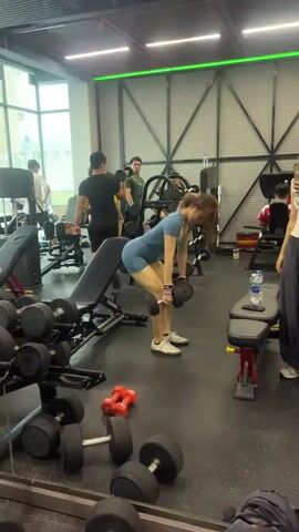 The video shows a woman in a gym performing dumbbell rows. She is wearing a blue athletic outfit and white sneakers. She is bent over at the hips, with her back straight, and is holding dumbbells in each hand. She is actively lifting the dumbbells towards her chest, engaging her back muscles. Other people are visible in the background, some are working out, others are standing. The environment is a typical gym with weight racks, benches, and other exercise equipment.