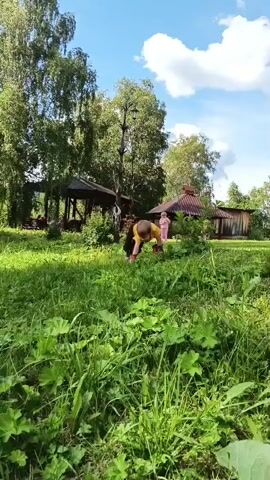 The media file captures a child playing in a grassy area on a sunny day. The child, wearing a yellow shirt and dark pants, is on their hands and knees, seemingly interacting with a red object on the ground. In the background, there are two gazebos and a wooden structure, suggesting a park or recreational area. Another person, dressed in pink, is visible standing near one of the gazebos. Tall trees with green foliage surround the scene, indicating a summer setting. The sky is blue with white, fluffy clouds, and the overall atmosphere appears relaxed and cheerful, with the bright sunlight illuminating the greenery. The time of day is likely midday or early afternoon, given the strong lighting. There is no visible text in the media file. The location context is Medvedevka, Russia.
