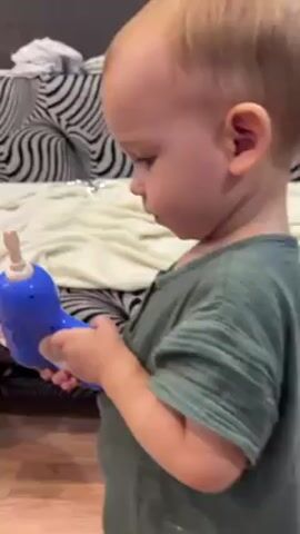The video shows a toddler and a young man. The toddler, who appears to be a boy, is holding a blue toy and appears to be trying to use it, seemingly as part of some sand-based play activity on the floor. The young man is shirtless and has a blue plastic toy hat on his head. The video is home footage and appears to be taken with a handheld device.