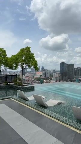 A 4K vertical video opens from a low angle showing a sleek infinity pool on a rooftop. White lounge chairs are partially submerged in the pool. In the background, a cityscape with numerous buildings of varying heights is visible under a cloudy sky. A single green tree stands to the left of the pool. The camera pans slowly to the right, revealing more of the city skyline and the expansive pool. The sound of gentle splashing water can be heard.