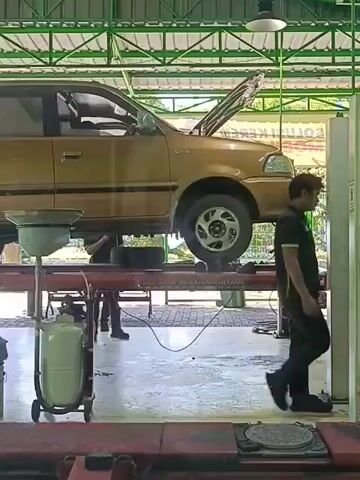 The video shows a gold-colored car being serviced in an auto repair shop. The car is raised on a hydraulic lift, with its hood open. A drain pan is positioned beneath the car, presumably to catch fluids. A person in dark clothing can be seen working on the car, partially obscured by the lift. Another person in dark clothing is visible in the background on the right side of the frame, walking away. The shop appears to be well-lit, with a green-colored ceiling structure and white walls. Various tools and equipment are visible in the background, including what looks like a large, green tank on wheels. The floor is concrete and appears somewhat wet or dirty, indicating an active work environment. The overall scene depicts a routine car maintenance or repair operation.
