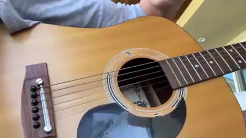 A person is playing an acoustic guitar. The guitar is a light brown color with a black pickguard. The person is wearing a white shirt and a silver bracelet. The person is strumming the guitar strings with their right hand. The scene appears to be indoors.