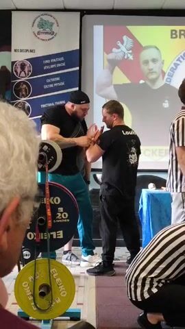 The video shows two men arm wrestling, with an official standing close to them in a striped shirt. Both men are in black shirts and are standing on a black, red and white checkered floor. One of the men is large, wearing a black cap, black shirt, turquoise pants, and white sneakers. He has tattoos on his right arm. The other man has lighter hair and is wearing black pants. To the left of them are multiple weight plates. In the background is a projector screen with a picture of a man holding up his arm. A poster with the words “FEDERATION FRANCAISE DE FORCE" is next to the screen. Part of the head of a white-haired man is visible in the left foreground. Another person is crouched in the right foreground. The event seems to be a part of a weightlifting competition.