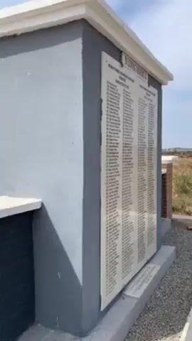 The video shows a gray and white cement structure with a commemorative plaque, bearing the inscription "In Loving Memory of..." and a list of names under the heading "Victims of 7th March, 2010 Massacre at Dogo Nahawa Plateau State Names of the Victims." The camera zooms in on the plaque, revealing more names and columns. The environment appears to be outdoors, possibly a memorial site or cemetery, with a backdrop of green vegetation and a partly cloudy sky. The video is presumably taken to record the memorial and the names of the victims listed.