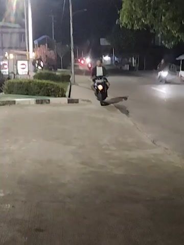 A person is riding a motorcycle at night. The person is wearing a light-colored shirt and dark shorts, with a dark jacket over the shirt. The motorcycle has a license plate that is partially visible. The person appears to be looking to their left. The background is dark with some lights from buildings and streetlights visible. There is a tree on the right side of the frame. The ground is paved. The camera is moving.