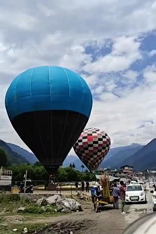 The video shows two hot air balloons in an outdoor setting during the daytime. The prominent balloon in the foreground is blue and black, while a smaller, checkered red, black, and white balloon is visible behind it to the right. Both balloons appear to be inflated and upright, possibly tethered or preparing for ascent.

Several people are scattered across the scene. Some individuals are near the base of the blue and black balloon, while others are closer to a dirt road on the right side of the frame. On this road, a white car and other vehicles are visible, suggesting traffic. A cart laden with what appears to be sugarcane is positioned near the road, with a few people standing around it.

In the background, lush green mountains rise under a sky with scattered white clouds. The overall lighting suggests it's a bright day. A notable detail is the license plate of the white car, partially visible as "HP 34D 832[5]", indicating an Indian state, likely Himachal Pradesh, which aligns with the mountainous landscape and the given location context of Jather, India. The scene captures a mix of recreational activity (hot air balloons) and everyday life (road traffic, street vendors).