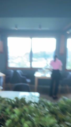 The video shows the inside of a restaurant with a scenic view of a town nestled in the mountains.

The interior of the restaurant features modern decor with gray and light-colored wooden furniture. Tables are set with glasses and cutlery. Greenery is placed as a decorative element on one of the tables in the foreground. A woman wearing a pink shirt and black pants is setting up a table near a large window.

The window offers a panoramic view of the town below, surrounded by tree-covered mountains in the background. The sky is bright, with some clouds visible. Several birds are seen flying in the distance.

A hand holding a white paper appears briefly at the bottom of the window frame.
