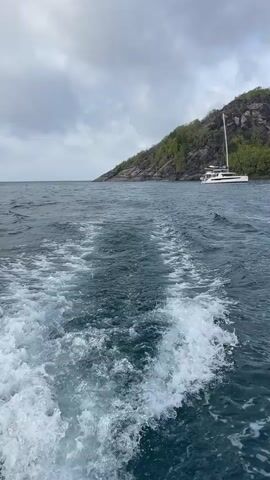 The video shows a choppy ocean with a white wake trailing behind the camera. In the distance, a white catamaran with two hulls and a tall mast is sailing on the ocean. Behind the catamaran is a large, green, tree-covered island with rocky terrain. The sky is cloudy and overcast. The video appears to have been filmed from another boat moving away from the catamaran and island.