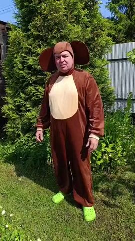 A person wearing a brown dog costume stands in a grassy yard. The costume has large floppy ears, a tan belly, and a tail. The person is wearing bright green Crocs. They are standing with their arms outstretched, palms facing outwards. Behind them is a large evergreen tree and a metal fence. The sun is shining, casting shadows on the grass. The person turns their body from facing the camera to facing left, then turns to face the back, and finally turns to face right.
