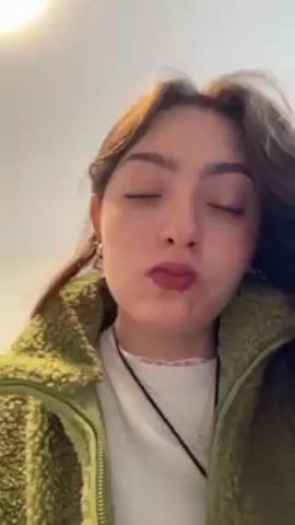 The video shows a young woman with light brown hair and green eyes. She is wearing a green fleece jacket and a white shirt with a black cord around her neck. She has a nose piercing and is wearing lip gloss. She holds a small, clear plastic bottle with a white applicator to her mouth, applies the product to her lower lip, and then closes the bottle. Afterward, the woman looks directly at the camera with her lips closed. The background is a plain, light-colored wall.