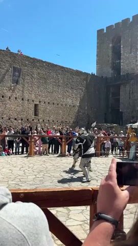 The video shows two people in full armor fighting with axes in a courtyard of a stone castle. A crowd is watching. A person in period clothes, possibly an emcee, is holding a yellow flag. A spectator is taking a video with a phone. The sky is clear and blue.