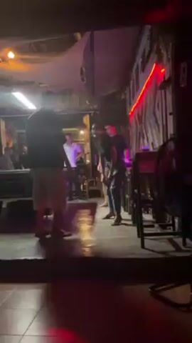Under the colorful lights of an outdoor bar, a woman and a man are dancing. The man wears a dark t-shirt and light-colored shorts, while the woman is in a black shirt and jeans. Behind them, a band is playing, with one member in a white shirt standing out. The bar's decor includes string lights, a corrugated metal wall, and wooden furniture, giving it a rustic vibe. Other people are seated at tables in the background, enjoying the music.