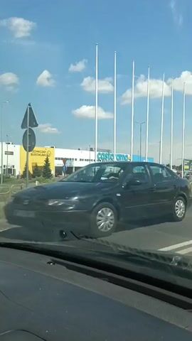 The video shows a view from inside a vehicle, looking forward and slightly to the right. The road ahead has white lane markings. To the left, there is a Castorama store with a large blue sign. Several tall, white flagpoles are visible in front of the store, with flags that are not clearly discernible. There are cars parked in the store's parking lot and some cars driving on the road. The sky is blue with scattered white clouds. A blue circular traffic sign with a white arrow pointing down and to the left is visible on a yellow pole in the foreground. Another similar yellow pole is present to the right of it. The video pans slightly to the right as it progresses.