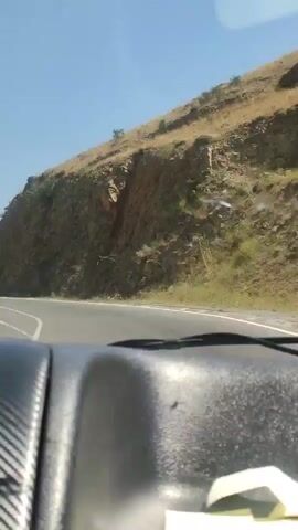 A first-person perspective video captured from inside a car shows a road winding through a mountainous landscape under a clear blue sky. The dashboard of the car is visible in the foreground, featuring a carbon fiber-like textured panel and air vents. A small, black helicopter figurine is placed on the dashboard, positioned above the air vents. The road ahead curves to the left, with a steep, rocky, and sparsely vegetated mountainside rising to the right. In the distance, the landscape continues with rolling hills, power lines, and what appears to be a tower structure on a hilltop. The car is in motion, suggesting a journey along this scenic route.