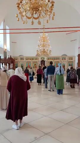 The video shows the interior of a church, with people standing or walking. The camera is positioned at a medium height, looking towards the altar area. There are large, ornate chandeliers hanging from the ceiling. The walls are white, and there are icons displayed on the walls and on an iconostasis. The floor is tiled in a checkerboard pattern. Several people are wearing headscarves or hijabs. The overall atmosphere appears to be one of a religious service or gathering.