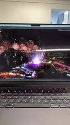 A person is watching a video on a laptop. The video shows a live performance with dancers on a field. The dancers are wearing red, white, and black costumes. There are stage lights and pyrotechnics. The laptop is open and on a table. The background is a blurry room.