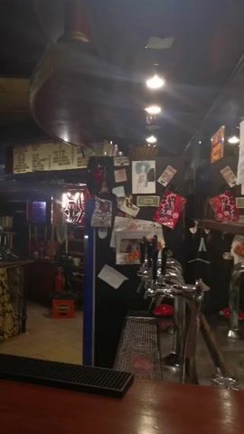 The video shows the interior of a bar. In the foreground, a wooden bar counter is visible with a black tray on it. Behind the counter, there are several beer taps. The background is cluttered with various items, including posters, pictures, and a fan. A menu board is displayed on the wall above the bar. In the left side of the frame, a person wearing an orange t-shirt can be seen behind the counter, appearing to be working. The lighting is dim, typical of a bar setting.