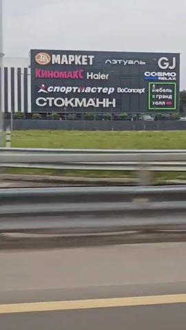 The video shows a view from a moving vehicle, looking towards a large commercial building complex with various signs. The signs indicate multiple businesses such as "МАРКЕТ", "Киномакс", "Haier", "Спортмастер", "ВоConcept", and "СТОКМАНН". Further along, other signs are visible including "Садовый Рай", "MR. DOORS", "М.видео", "DOM", "лента", and "ПЯТЕРОЧКА". The building has a modern architectural style with black and white vertical stripes. The environment appears to be an overcast day, with some greenery visible in front of the building and a highway barrier in the foreground. The camera perspective suggests the viewer is on an elevated road or overpass, looking down slightly at the complex. There are no people clearly visible in the footage. The movement of the vehicle causes a slight blur, particularly in the background.