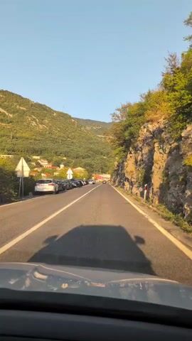 This is a first-person point of view video taken from inside a car traveling on a two-lane road. The car is moving forward, and the road is flanked by a steep, rocky hillside on the right and a line of parked cars and a village on the left. The sun is shining brightly, casting a shadow of the car on the road. The sky is clear and blue, and the surrounding landscape is mountainous and covered in greenery. There are no visible people in the immediate vicinity, and no specific actions or interactions are taking place beyond the car's movement and the general setting. The overall impression is that of a scenic drive through a coastal or mountainous region.