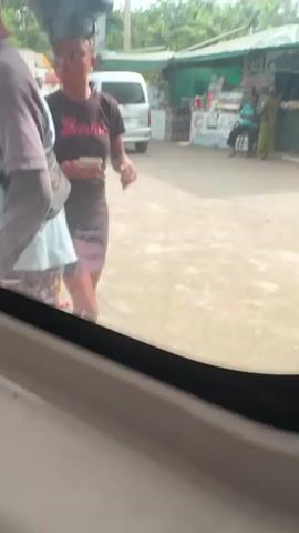 The video shows an outdoor scene in what appears to be a roadside area. A woman in a black dress and a "Barbie" t-shirt is walking along the road, holding a phone, with what looks like a bucket or container balanced on her head. There are other people around, including a man carrying a bag filled with what looks like dried banana chips. In the background, there's a small roadside stand or shop with the sign "BARBECUE & CHIPS" visible, as well as other merchandise on display. The ground is concrete, and there are trees and utility poles visible in the background, suggesting a semi-urban or rural setting.