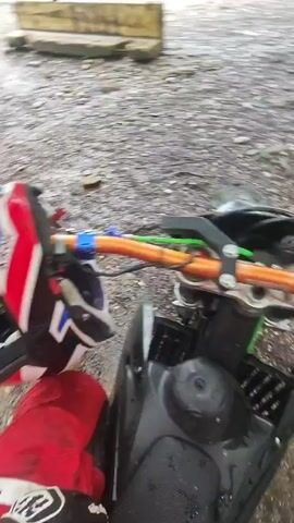 The video shows a person on a motorcycle, wearing a red and white motocross suit and an Airoh helmet. The motorcycle is parked on a muddy ground near a riverbed. The riverbed is wide and rocky, and the water is shallow and murky. The river is surrounded by green trees and vegetation. The camera pans across the scene, showing the river and the surrounding forest. The tone of the video is peaceful and serene.