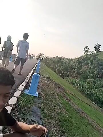 The video captures an outdoor scene along an elevated pathway, likely a dam or levee, under a bright, diffused sky. In the immediate foreground, a young male with dark, curly hair is visible from the chest up, looking intently at a smartphone held in his hands. He wears a dark t-shirt with some white, unreadable text (possibly "NORTHERN" or "NORTH") on it.

Behind him, three individuals walk away from the camera along a paved path. The person on the left is carrying a small child on their back or hip, obscured by the foreground subject. The middle person wears light-colored clothing, and the person on the right is dressed in a light t-shirt and shorts.

The pathway is lined on one side by light blue, cylindrical bollards or markers. To the right of the path, there's a sloping grassy embankment covered in green foliage and distant trees. The left side of the path, where the foreground subject is positioned, appears to border a body of water, though it's mostly out of frame.

The overall lighting suggests either early morning or late afternoon, with a clear, hazy sky and soft, even illumination. The weather appears calm. No specific emotions are discernible from the distant walkers, and the foreground subject is focused on their device. The scene conveys a tranquil, recreational atmosphere in an open, natural setting, consistent with the Jambeyan, Indonesia context.