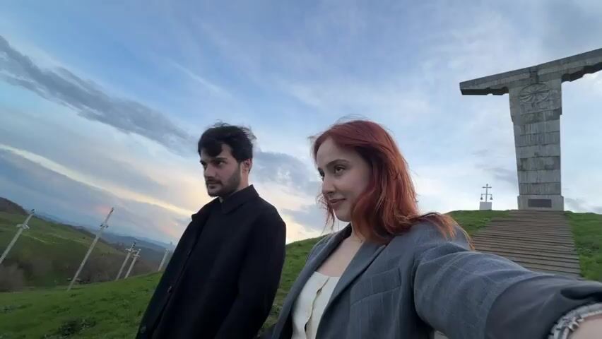 A young couple stands on a grassy hill.  The woman, with auburn hair and a gray blazer, holds the camera and smiles. The man, with dark hair and a black coat, stands beside her.  Behind them are a set of concrete stairs leading up to a large concrete monument and several swords are placed in the ground on the grassy hill. The man touches the woman's face and kisses her cheek. The sky is a mix of blue and gray clouds, indicating early evening.
