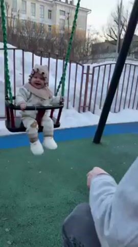 The video shows a baby being pushed on a swing outdoors, on a partly sunny day. The baby is wearing a beige bonnet, bib, and matching outfit with white booties. The baby is laughing and appears to be enjoying the ride. The swing is red and black, with green ropes. An adult wearing a grey sweatshirt is pushing the swing. The ground is a mix of green and blue playground flooring, and there is snow piled up near a white fence in the background. There are buildings visible in the distance.