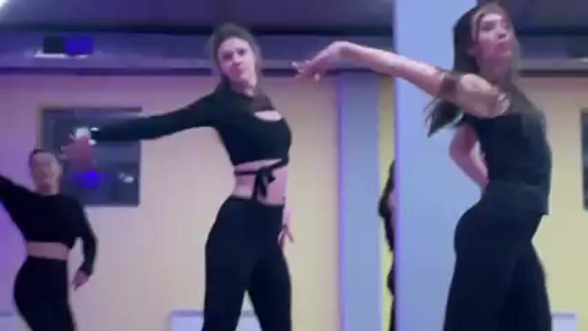 Two women are dancing in a dance studio, both wearing black outfits. The woman on the left is wearing a long-sleeved crop top and pants, while the woman on the right is wearing a tank top and pants. They are facing the camera and performing a choreographed dance routine. There are two other women in the background, one partially obscured by a pillar, who appear to be part of the same dance group. The studio has yellow walls with a window and is lit with bright overhead lights and pink accents.