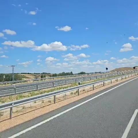 The video shows a view from a vehicle traveling on a highway. The sky is blue with scattered white clouds. The highway has multiple lanes separated by metal guardrails. On the left side of the highway, there are fields and some trees. On the right side, there is a sloping embankment covered in dry grass and some sparse vegetation. Several vehicles, including trucks and cars, are visible on the highway in the distance. The sun appears to be high in the sky, indicating daytime.