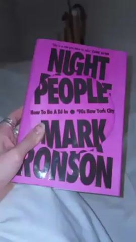 A hand holds a pink book titled "Night People: How To Be A DJ In '90s New York City" by Mark Ronson. The book is shown on top of a white bedsheet. The lighting is dim.