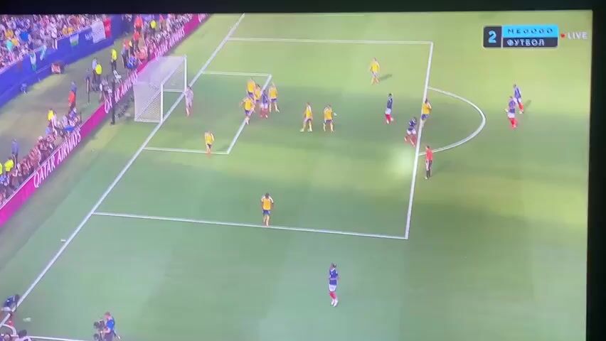 An aerial view of a soccer match shows players in yellow and blue uniforms on a green field. The game is being broadcast on a screen, with a score graphic in the upper right corner displaying "2" and "МЕГООО ФУТБОЛ LIVE". The camera focuses on a play near the goal, where players are actively engaged. A player in a yellow uniform is dribbling the ball towards the goal. Another player in a blue uniform is attempting to defend. Several other players from both teams are positioned around the penalty area, observing the action and preparing to react. The background shows a stadium with spectators and advertising boards.