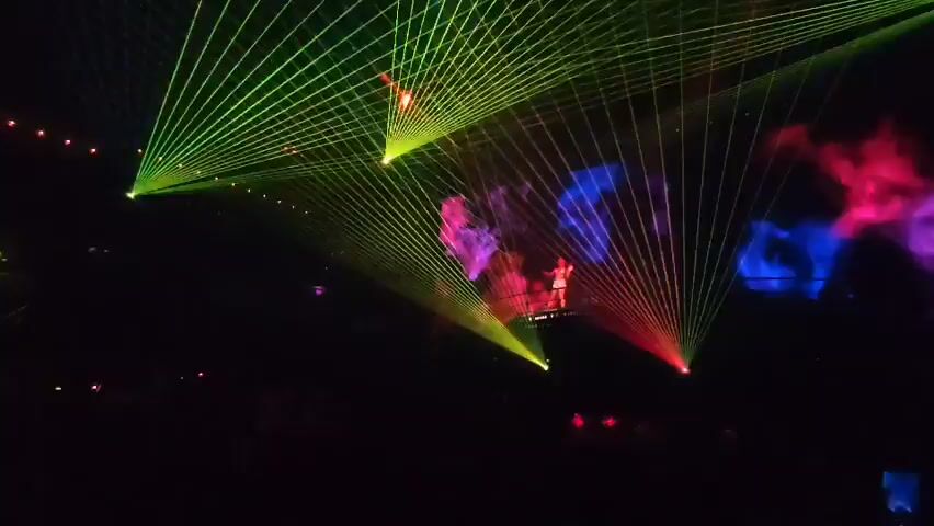 The video shows a crowded concert venue bathed in bright, colorful lights. The crowd appears to be enjoying the show, with many people raising their hands. A performer stands on a raised platform, silhouetted against the lights. The lighting is dynamic, with beams of light scanning the room and changing colors. The overall atmosphere is energetic and festive.