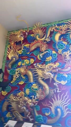 This is a vertical, eye-level, medium shot video of an ornate wall decoration featuring multiple golden dragons in various dynamic poses. The dragons are intricately detailed with scales, claws, and flowing manes, some appearing to be roaring or in mid-flight. They are set against a deep red background and surrounded by stylized, puffy clouds in shades of blue, green, and yellow, giving the impression of a celestial or mythical scene. The artwork is highly three-dimensional, with elements protruding from the wall. Towards the bottom of the frame, a doorway or archway is visible, allowing a glimpse of an outdoor scene with lush green foliage and bright sunlight. The entrance is flanked by more decorative elements, including what appears to be a traditional Chinese-style latticed door with a yin and yang symbol. The overall impression is one of grandeur and cultural significance, likely depicting a temple or traditional building interior.