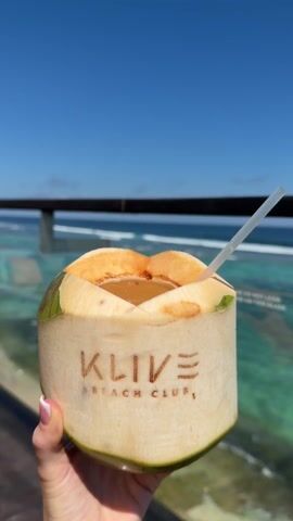 The video shows a person holding a coconut drink with a straw on a sunny day. The coconut is labeled "KLIVE BEACH CLUB" and is held up against a backdrop of a beautiful beach and clear blue ocean. The water is turquoise near the shore and becomes a deeper blue further out.  A black railing and glass panel are visible in the foreground, with the text "PLEASE DO NOT LEAN OR CLIMB ON THIS GLASS" printed on the panel. The camera pans slightly upward to show the horizon line and the clear blue sky.