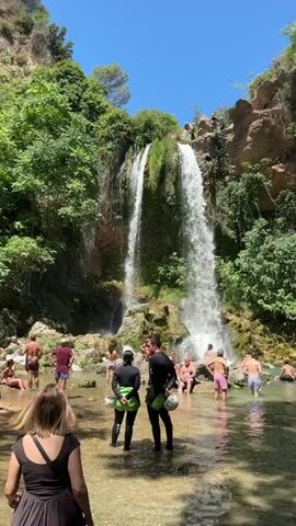 A vertical, outdoor shot shows a group of people gathered at the base of a tall, multi-tiered waterfall. The water cascades down a rocky, vegetation-covered cliff face into a clear, shallow pool. The day is bright and sunny, with a clear blue sky overhead. Several people are in the water, some wading, others sitting or standing. Two individuals in the foreground are wearing wetsuits and appear to be preparing for an activity. Other individuals are dressed in swimwear and casual clothing, standing or sitting on the rocks and the water's edge. The surrounding environment is lush with green trees and foliage. The scene captures a lively atmosphere of people enjoying a natural recreational area.