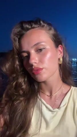 The video begins with a close-up of a young woman's face, her hair flowing around her. She is wearing a light-colored top and gold jewelry. The background is dark, with blurred lights suggesting a nighttime setting, possibly on a boat or near a body of water. The woman looks directly at the camera, her mouth slightly open. The scene then shifts to a view of choppy, dark water, possibly the ocean, with a pink and cream-colored edge visible in the foreground. The water is illuminated by faint light, creating a textured surface. The video then returns to the woman's face, but this time she appears to be in a different setting, with another person's blurred blonde hair visible next to her. The background remains dark with distant lights. The woman’s expression changes to one of surprise or excitement, her eyes wide and mouth open. The final shot is a very blurry image of the woman's face with her mouth wide open, her expression animated.