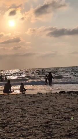 The video shows a beach scene at sunset. The sun is visible in the upper left corner, casting a warm glow on the water and clouds. Several people are present, some sitting on the sand and others in the shallow water. In the foreground, the sand is textured and appears damp near the water's edge. Waves are gently rolling onto the shore, creating a serene atmosphere. The overall impression is one of a peaceful evening at the beach.