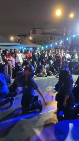 A group of young people in matching uniforms are performing a synchronized dance routine outdoors at night. They are on a paved surface that is illuminated by stage lighting, including purple and white lights. A crowd of onlookers stands behind a rope barrier, watching the performance. String lights and streetlights are visible in the background, along with some tents and buildings. The dancers are on their knees and in various dynamic poses, with some members reaching their arms up and others looking towards the sky. The performance appears to be energetic and engaging, with the dancers moving in unison.