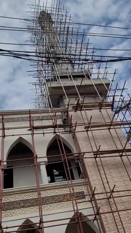 The video shows a mosque undergoing construction or renovation, as evidenced by the scaffolding surrounding the building. The mosque is painted white, with ornate decorations, including Arabic calligraphy. The architecture features arches on the facade, and there is a minaret covered in scaffolding as well. Workers can be seen on top of the structure. The sky is visible in the background, with some clouds.
