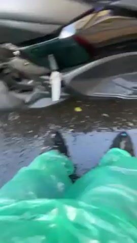 The video shows a close-up view of a parked grey Yamaha NMAX scooter with a black seat. The scooter is positioned next to a yellow vehicle, possibly a taxi, with the rear of the vehicle visible behind the scooter. The ground appears wet, possibly from rain, and some leaves are scattered around the scooter. The camera pans around the scooter, showing details such as the handlebars, seat, exhaust pipe, and the "NMAX" logo. A helmet is sitting on the scooter's handlebars.