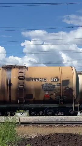 This is a side-view, medium shot video of a moving train carrying multiple tank cars. The train is on a railway track that runs horizontally across the frame. The sky above is blue with fluffy white clouds. In the foreground, there is lush green vegetation on the left and brown soil on the right. The tank cars are large, cylindrical containers, some appearing to be made of metal and others painted in different colors, such as blue, light brown, and beige. Several of the tank cars have visible markings and numbers, including "58273566," "501 93572," "51760593," "500 80787," and "539 05576." There are also ladders attached to the sides of some of the tank cars. On the right side of the frame, a person's head and shoulders are visible as they are holding a smartphone, presumably filming or photographing the train. The train appears to be in motion, indicated by the slight blur on the wheels and the continuous movement of the tank cars across the screen.